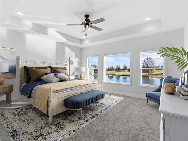 carpeted bedroom featuring baseboards, a water view, a tray ceiling, recessed lighting, and a ceiling fan