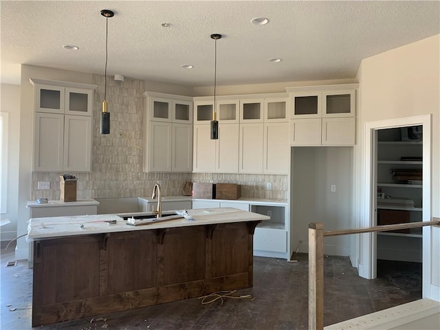 kitchen with a sink, glass insert cabinets, unfinished concrete flooring, a textured ceiling, and a kitchen island with sink