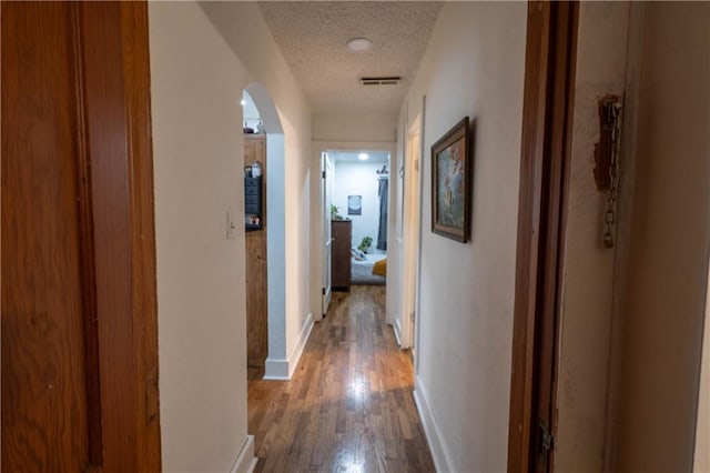 corridor with arched walkways, visible vents, a textured ceiling, wood finished floors, and baseboards