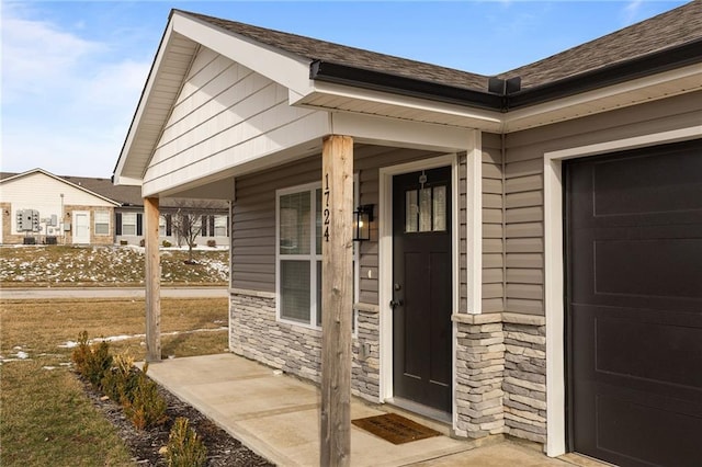 doorway to property featuring an attached garage, stone siding, a shingled roof, and a porch