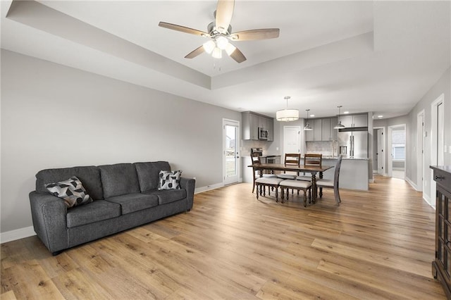 living area featuring a tray ceiling, baseboards, ceiling fan, and light wood finished floors