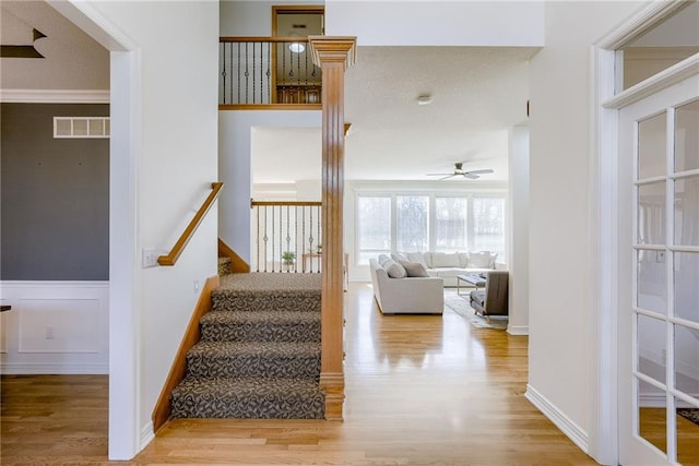 stairway with a wainscoted wall, crown molding, visible vents, ceiling fan, and wood finished floors