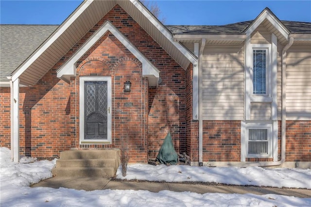 snow covered property entrance with brick siding and roof with shingles