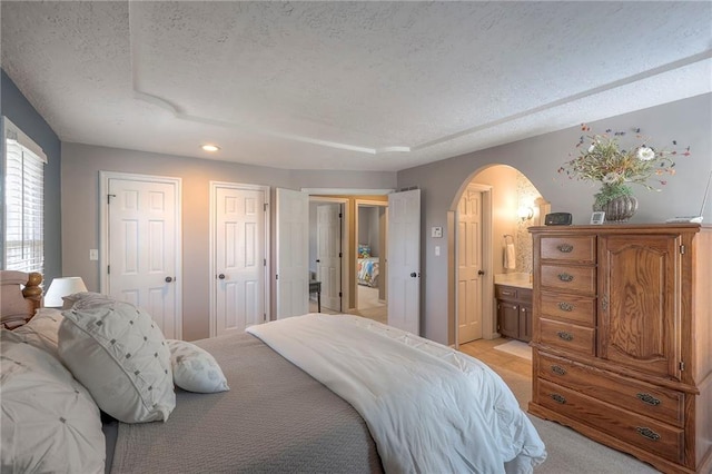 bedroom featuring arched walkways, a textured ceiling, ensuite bathroom, light colored carpet, and two closets