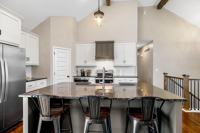 kitchen with beamed ceiling, appliances with stainless steel finishes, an island with sink, and dark wood-type flooring