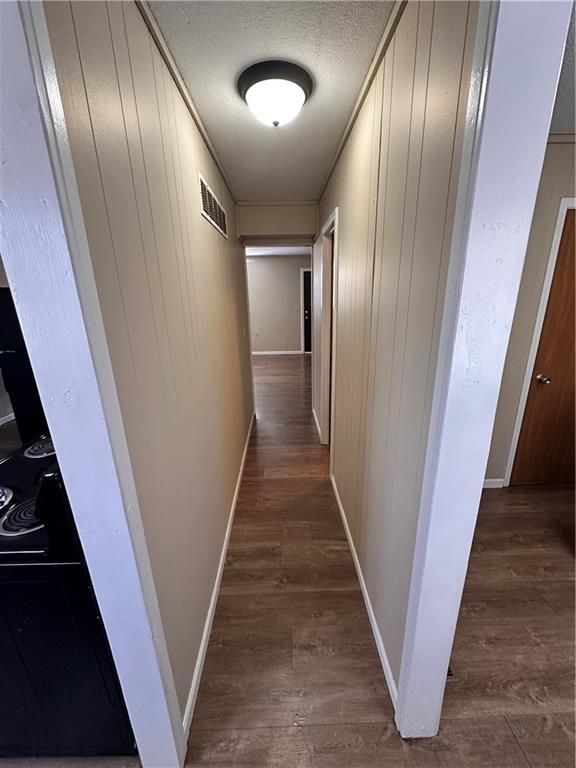 hall with a textured ceiling, wood walls, visible vents, baseboards, and dark wood finished floors