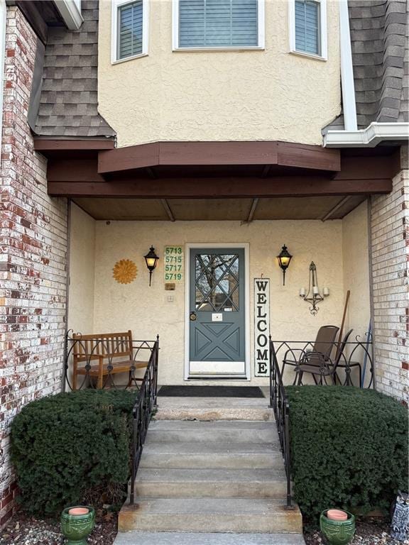 doorway to property with brick siding, stucco siding, a porch, and a shingled roof