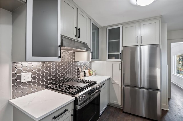 kitchen featuring decorative backsplash, under cabinet range hood, light stone counters, appliances with stainless steel finishes, and glass insert cabinets