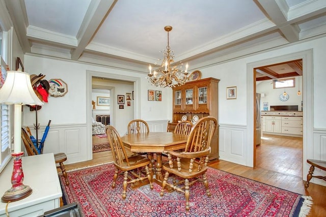 dining area with a wainscoted wall, light wood-style flooring, crown molding, beamed ceiling, and a notable chandelier