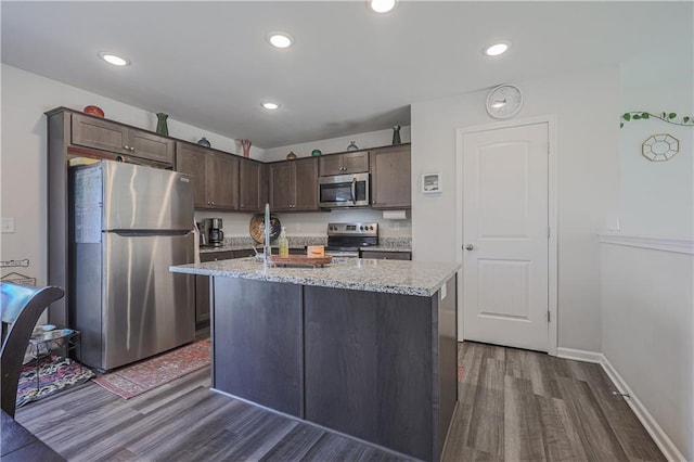 kitchen with dark wood-type flooring, light stone countertops, dark brown cabinetry, appliances with stainless steel finishes, and a kitchen island with sink