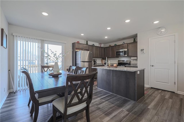 kitchen with a kitchen island with sink, stainless steel appliances, baseboards, dark brown cabinets, and dark wood-style flooring