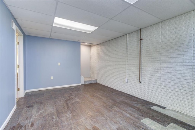 finished below grade area with brick wall, baseboards, a drop ceiling, and wood finished floors