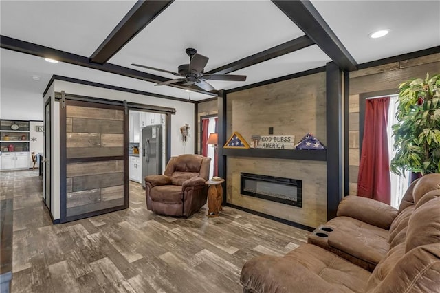 living area featuring ceiling fan, a barn door, wood finished floors, beam ceiling, and a glass covered fireplace