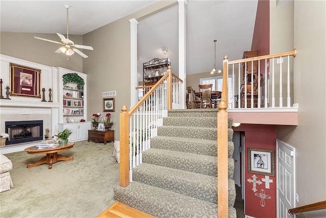 stairway featuring visible vents, carpet floors, high vaulted ceiling, a fireplace with raised hearth, and ceiling fan with notable chandelier