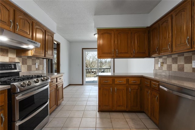 kitchen with light tile patterned floors, brown cabinetry, a peninsula, stainless steel appliances, and under cabinet range hood