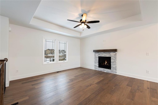 unfurnished living room with baseboards, dark wood finished floors, ceiling fan, a stone fireplace, and a raised ceiling