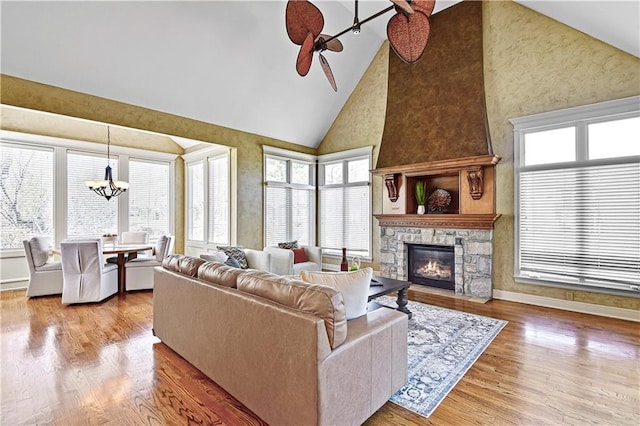 living room with high vaulted ceiling, ceiling fan with notable chandelier, wood finished floors, a stone fireplace, and baseboards