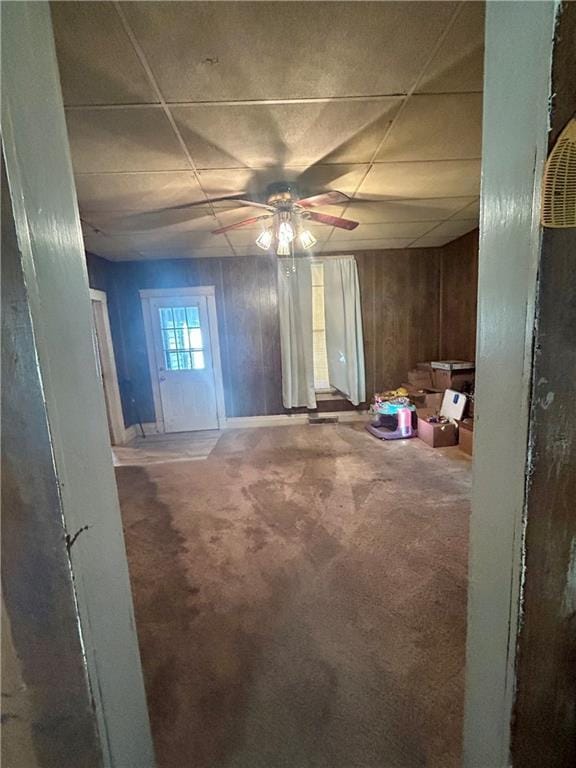 interior space with a drop ceiling, carpet, ceiling fan, and wood walls