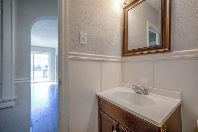 half bathroom with vanity, wainscoting, a textured wall, a decorative wall, and a textured ceiling