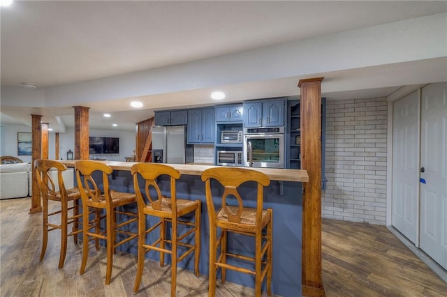 kitchen featuring blue cabinetry, wood finished floors, stainless steel appliances, a breakfast bar area, and brick wall