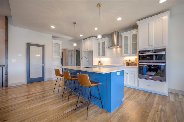 kitchen with a sink, double oven, light wood-style flooring, wall chimney exhaust hood, and a kitchen island with sink