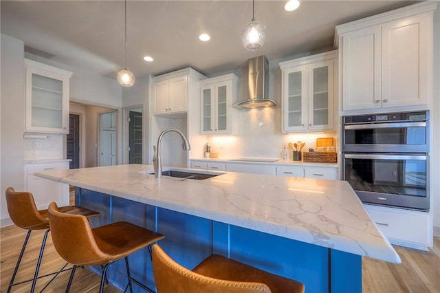 kitchen with a sink, white cabinets, light wood-style floors, double oven, and wall chimney range hood