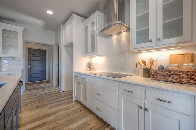 kitchen featuring light wood-type flooring, light stone counters, wall chimney exhaust hood, white cabinets, and black electric stovetop