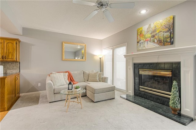 living area featuring a ceiling fan, a tiled fireplace, a textured ceiling, baseboards, and light colored carpet