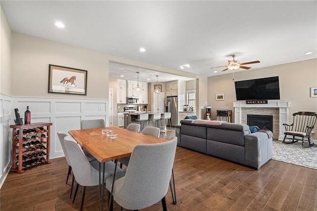 dining space with a decorative wall, ceiling fan, dark wood finished floors, a stone fireplace, and a wainscoted wall