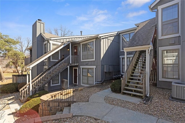 rear view of property with board and batten siding, stairs, roof with shingles, central AC unit, and a chimney