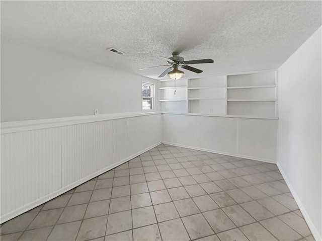 empty room featuring visible vents, built in shelves, a wainscoted wall, a textured ceiling, and a ceiling fan