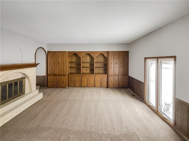 unfurnished living room featuring a brick fireplace, light carpet, wood walls, and wainscoting