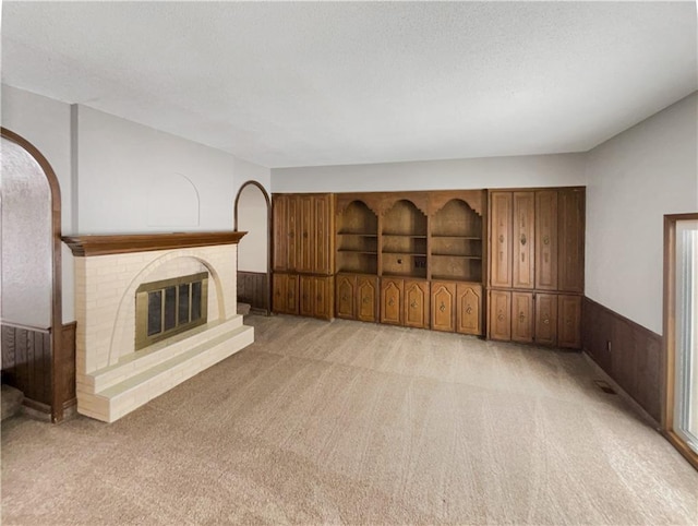 unfurnished living room with a textured ceiling, a brick fireplace, a wainscoted wall, and light carpet