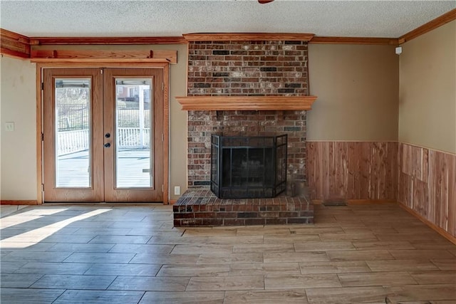 unfurnished living room with a textured ceiling, a fireplace, ornamental molding, french doors, and wainscoting