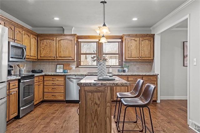 kitchen with a kitchen island, crown molding, dark wood-type flooring, appliances with stainless steel finishes, and brown cabinetry