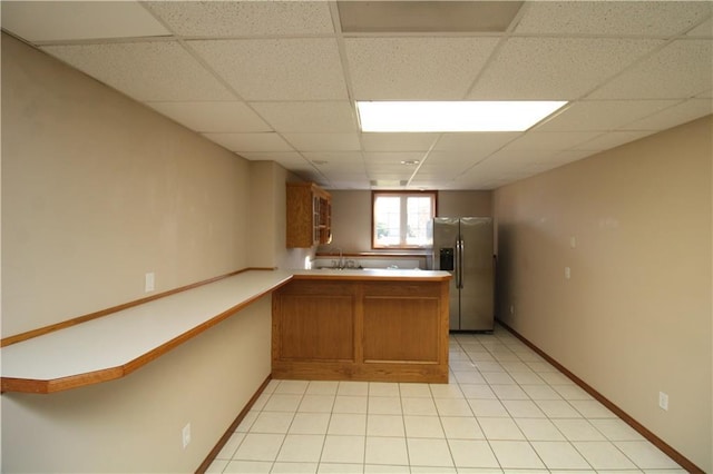 kitchen with brown cabinets, a drop ceiling, a peninsula, stainless steel fridge with ice dispenser, and light tile patterned floors