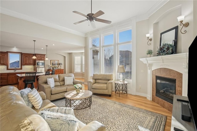 living room featuring light wood finished floors, ceiling fan, baseboards, a tiled fireplace, and ornamental molding