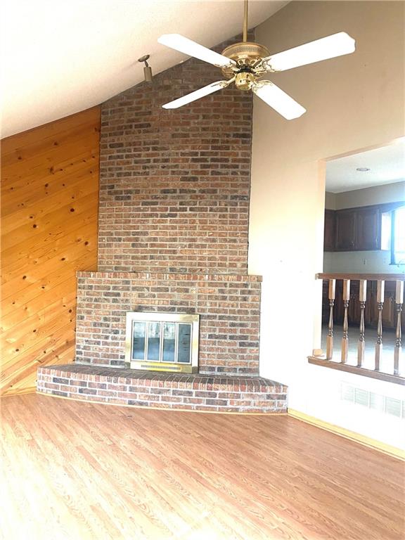 unfurnished living room featuring a brick fireplace, wood walls, wood finished floors, high vaulted ceiling, and a ceiling fan