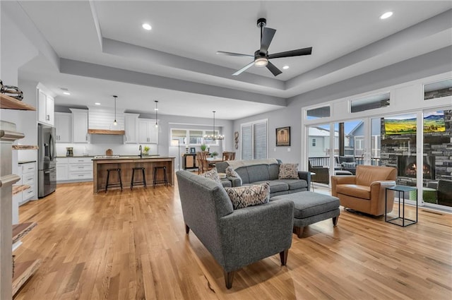 living room featuring ceiling fan with notable chandelier, a raised ceiling, light wood-style flooring, and recessed lighting