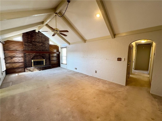 unfurnished living room with a brick fireplace, ceiling fan, light colored carpet, lofted ceiling with beams, and arched walkways