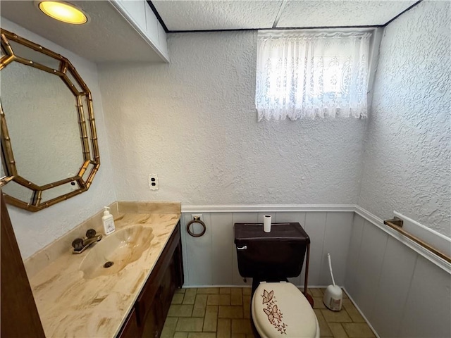 bathroom featuring vanity, toilet, a textured wall, and a wainscoted wall