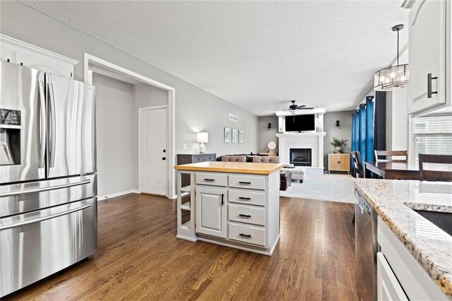 kitchen with a fireplace with raised hearth, ceiling fan with notable chandelier, stainless steel appliances, dark wood-style floors, and white cabinetry