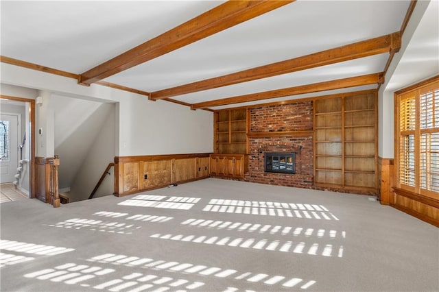 unfurnished living room featuring beamed ceiling, built in shelves, carpet, and wainscoting