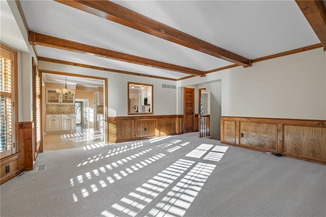 carpeted empty room with an inviting chandelier, beamed ceiling, visible vents, and wainscoting