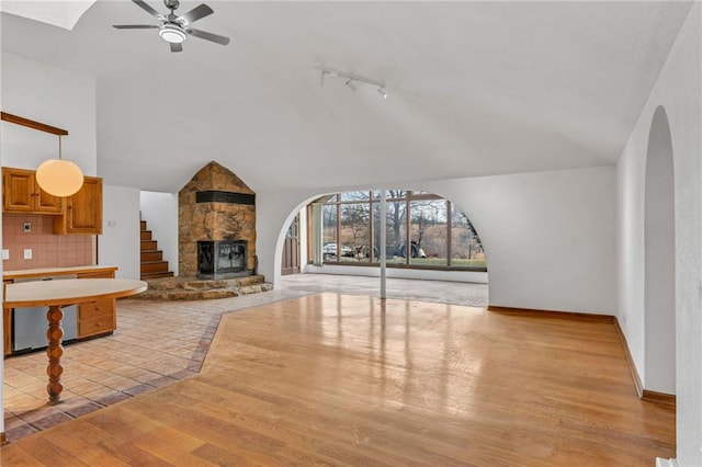 living room featuring a ceiling fan, light wood finished floors, arched walkways, a stone fireplace, and vaulted ceiling