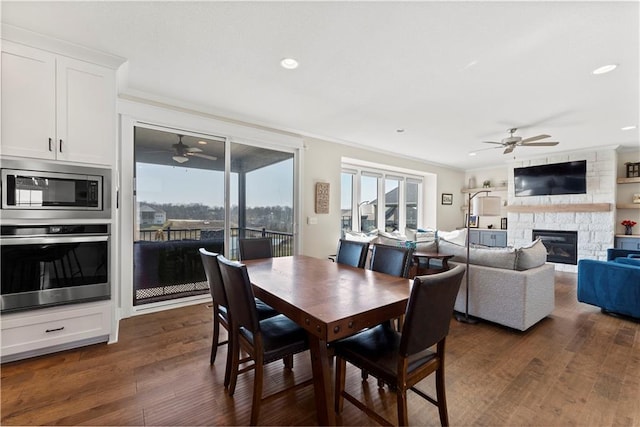 dining room with ceiling fan, ornamental molding, a stone fireplace, recessed lighting, and dark wood-style flooring