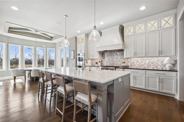 16121 Paradise St, Overland Park, KS 66221 - kitchen with premium range hood, backsplash, an island with sink, a tray ceiling, and dark wood-style floors