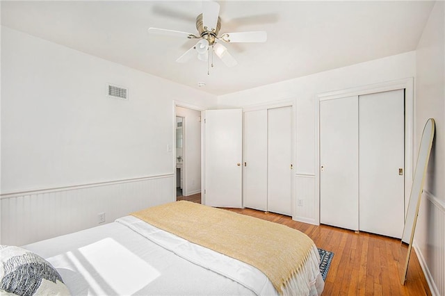 bedroom with a ceiling fan, visible vents, wood-type flooring, wainscoting, and two closets