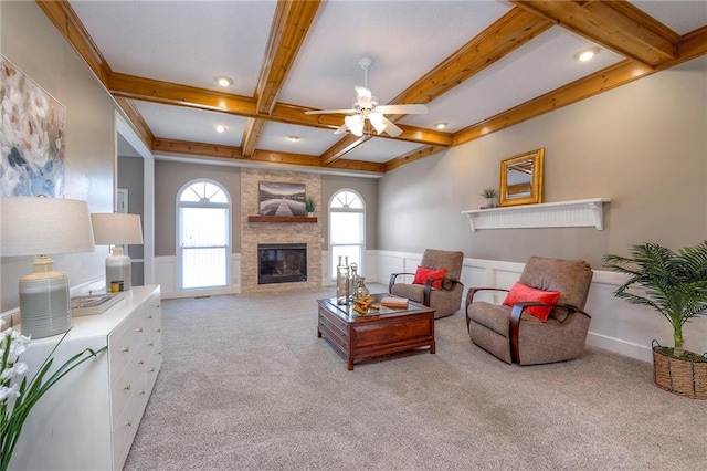 living area featuring a ceiling fan, a wainscoted wall, beam ceiling, a fireplace, and light colored carpet