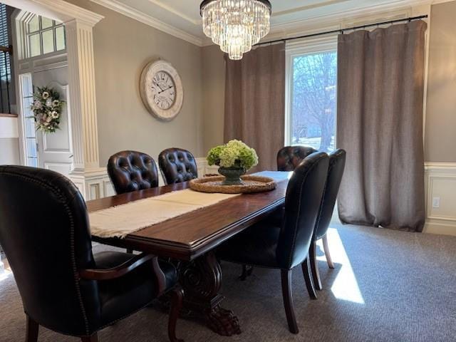 carpeted dining space with crown molding, a chandelier, wainscoting, a decorative wall, and ornate columns